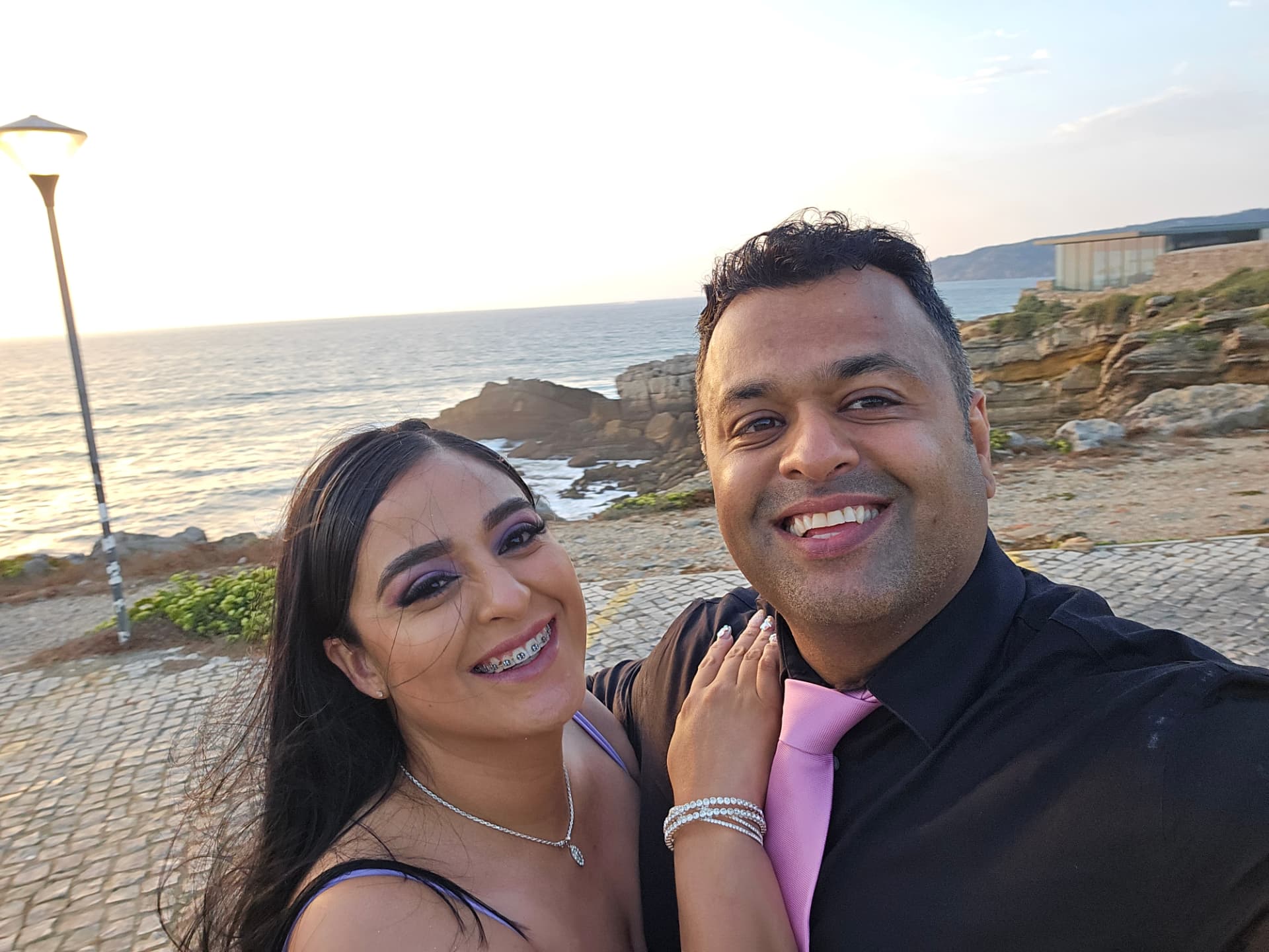Margarita and Samir taking a selfie on a cobblestone walkway by the sea at sunset